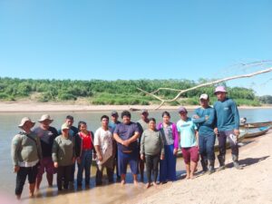 Foto de um grupo de indígenas Manchineri sobre uma praia de areias brancas na margem de um igarapé com o céu azul ao fundo.