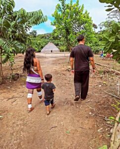 Clayton Rodrigues caminha ao lado de uma indígena Marubo e seu filho dentro de uma aldeia. Ao fundo se vê uma casa tradicional Marubo.
