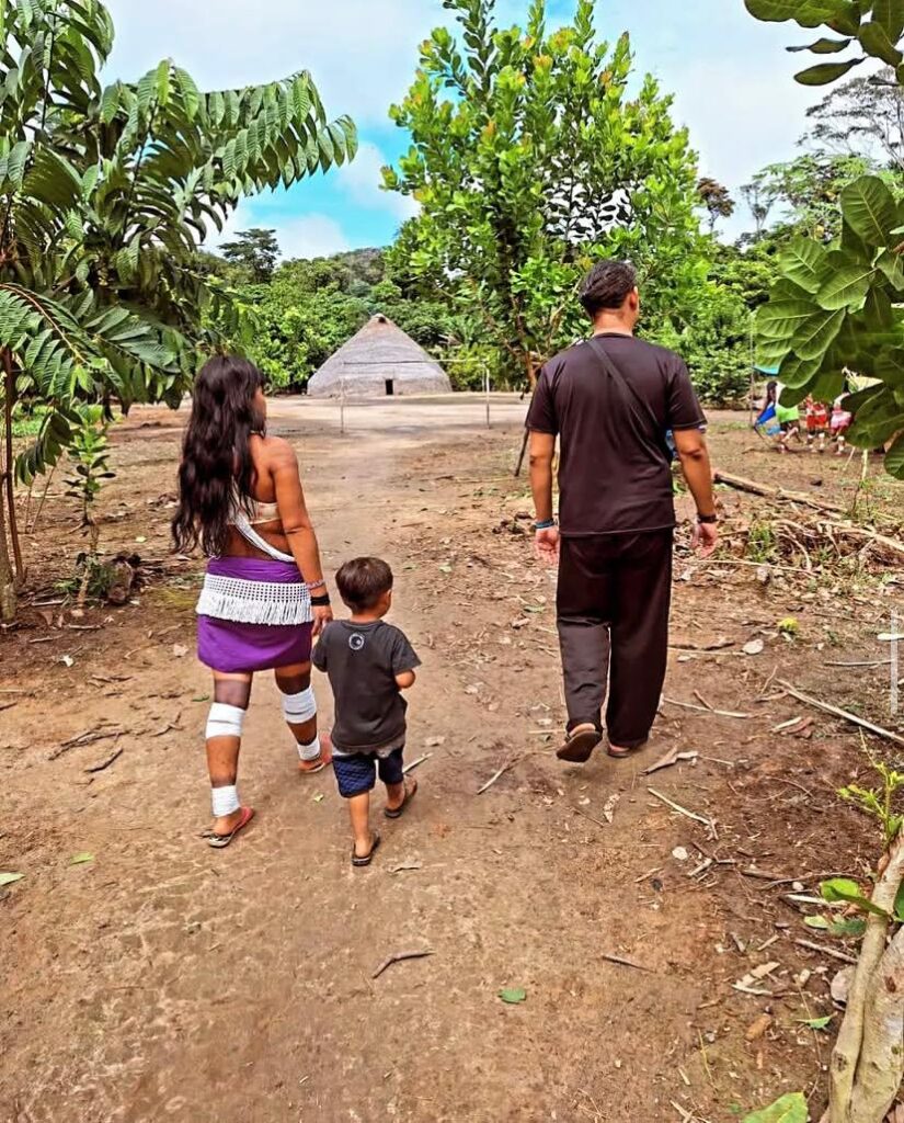 Clayton Rodrigues caminha ao lado de uma indígena Marubo e seu filho dentro de uma aldeia. Ao fundo se vê uma casa tradicional Marubo.